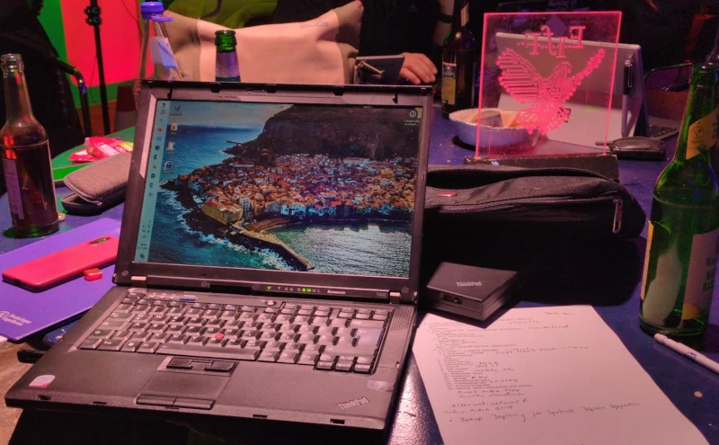 Ein offener Laptop auf einem Tisch. Daneben Flaschen, die FIfF-Taube als Plexiglasaufsteller und ein Blatt Papier mit einer Liste benötigter Software.