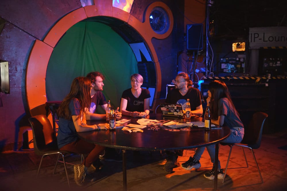 Innenansicht der c-base in Berlin, einem Hackerspace mit futuristischem, technikgeprägtem Design. Der Raum ist mit Bildschirmen, Kabeln und leuchtenden Elementen ausgestattet, was an das Innere eines Raumschiffs erinnert. Eine gruppe diverser Menschen diskutiert an einem Runden Tisch.