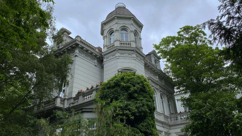 Die weiße Jugendstil Villa ist bewachsen mit Efeu. Grüne Bäume säumen das Gebäude.